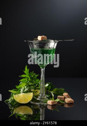 Glass of absinthe with ingredients on a black background. Absinthe spoon, brown sugar, sagebrush branch, and lime slice. Stock Photo