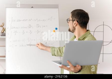 Young male math teacher with laptop and formulas on whiteboard giving online lesson at home Stock Photo