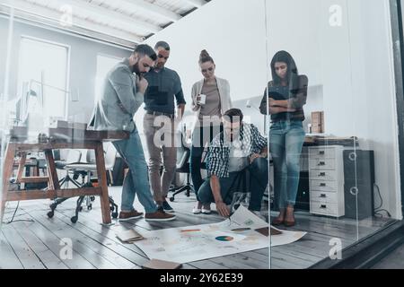 Discussing strategy. Handsome young man with beard pointing at large paper laying on floor while his colleagues looking at it Stock Photo