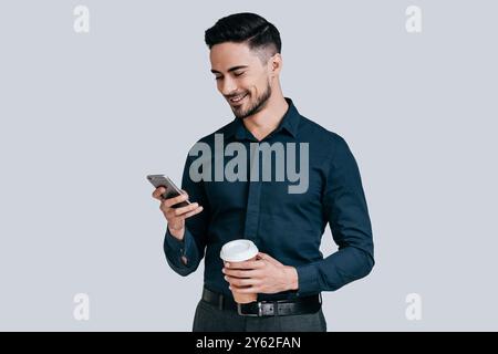 Nice chat. Good looking young man holding smart phone and looking at it with smile while standing against grey background Stock Photo