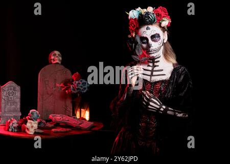 Portrait of woman holding black rose set against dark background grave site celebrating Dia de los Muertos. Eerie lady with flower surrounded by creepy funeral items during Halloween horror event Stock Photo
