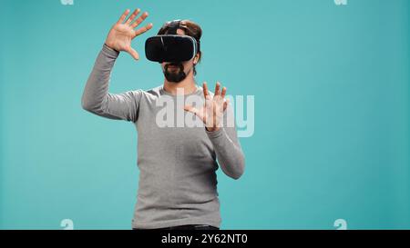 Man wearing virtual reality headset, interacting with interface, doing swiping motions. Person having fun using high tech futuristic modern VR gadget, isolated over blue studio background, camera B Stock Photo