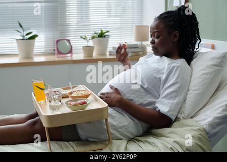 Pregnant woman sitting on hospital bed and eating meal while holding a piece of food, surrounded by a tray with various dishes and beverages, with plants on the windowsill Stock Photo