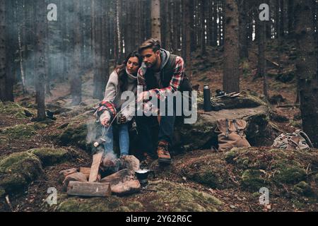 Enjoying time together. Beautiful young couple roasting marshmallows over a campfire while sitting in the forest Stock Photo