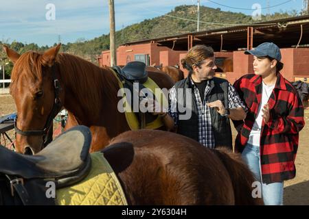 Two skilled man woman stable workers in plaid shirts standing close to saddled horse and discussing upcoming work in countryside club Stock Photo