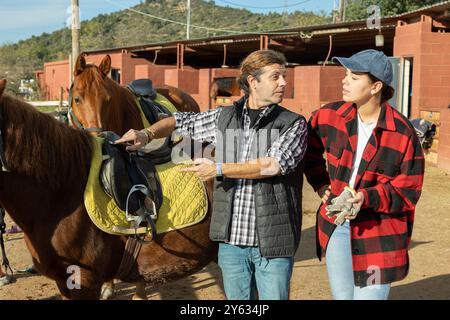 Two skilled man woman stable workers in plaid shirts standing close to saddled horse and discussing upcoming work in countryside club Stock Photo