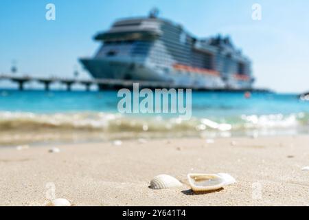 Seashells rest on a sandy beach, with a cruise ship blurred in the background, creating a serene and tranquil scene. Essence of a relaxing vacation. Stock Photo