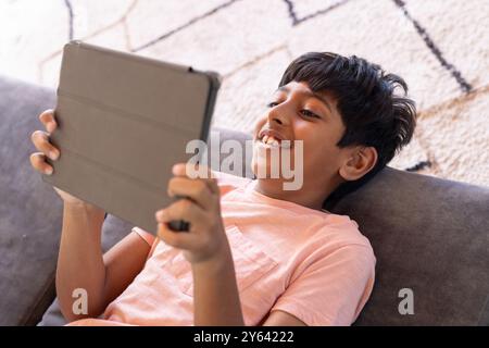 Smiling Indian boy using tablet while lying on couch at home, enjoying screen time Stock Photo