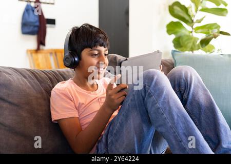 Wearing headphones, Indian boy sitting on couch using tablet and smiling Stock Photo