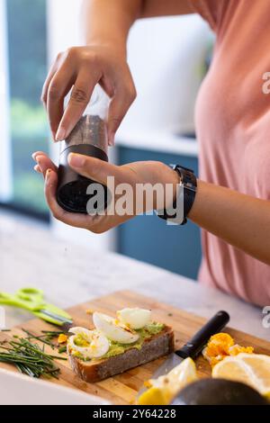 Seasoning avocado toast with pepper, woman preparing healthy breakfast in kitchen, at home, at home Stock Photo