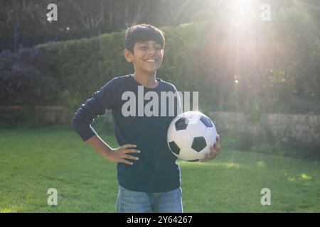 Smiling Indian boy holding soccer ball, standing in sunny garden, enjoying outdoor activity Stock Photo