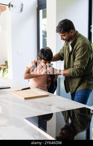 Helping son with backpack, indian father preparing him for school in kitchen Stock Photo