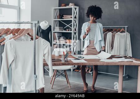 Generating fresh ideas. Full length of young African woman touching lips with pencil and looking at paper while standing in her workshop near the clot Stock Photo