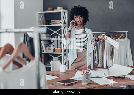 Searching for right answer. Thoughtful young African woman talking on smart phone and holding a paper while standing in her workshop Stock Photo