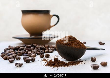 Freshly ground aromatic coffee in a measuring spoon against a background of scattered coffee beans and a ceramic cup and saucer in the background Stock Photo