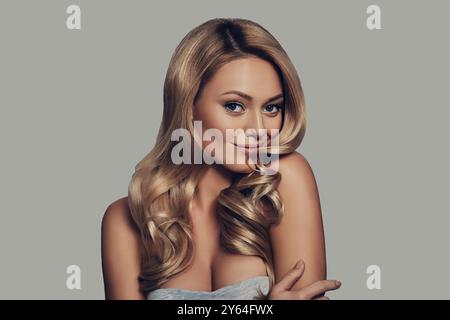 Simply stunning. Attractive young woman with long curly blond hair smiling and looking at camera while standing against grey background Stock Photo