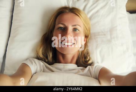 Close up portrait of happy smiling young woman stretching her hands for taking selfie with smartphone while lying on the bed in bedroom at home Stock Photo