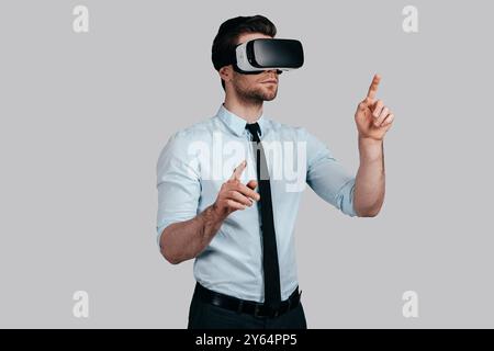 Experiencing virtual reality. Handsome young man in formalwear wearing virtual reality headset and gesturing while standing against grey background Stock Photo