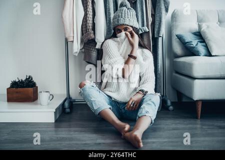 Trying to hide. Playful young woman in casual wear and knit hat covering face with her sweater while sitting on the floor with clothes hanging in the Stock Photo