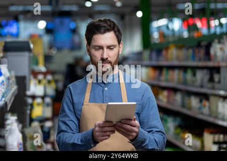 Grocery store worker in apron using digital tablet to manage inventory. Employee stands in supermarket aisle, focused on device, with shelves of products in background Stock Photo
