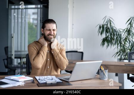 Businessman feeling stressed experiencing neck pain at workplace. Male office worker in office using laptop, expressing discomfort. Concept of work stress, health issues, and ergonomic challenges. Stock Photo