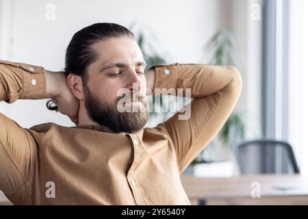 Bearded male office worker relaxing in contemporary office setting. Person enjoying a serene moment leaning back in chair, hands behind head, with closed eyes. Stock Photo