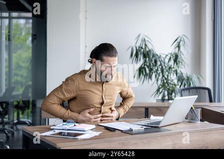 Businessman suffering from stomach pain in modern office environment, seated at desk with laptop and paperwork. Stressful work scenario depicted indoors with an emphasis on discomfort Stock Photo