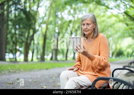 Older woman seated on park bench using tablet, engaging with digital technology. Outdoor setting emphasizes tranquil lifestyle, blending modern technology with nature. Stock Photo