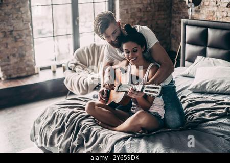 You are playing great! Handsome young bearded man teaching his girlfriend to play guitar while both sitting in bed at home together Stock Photo