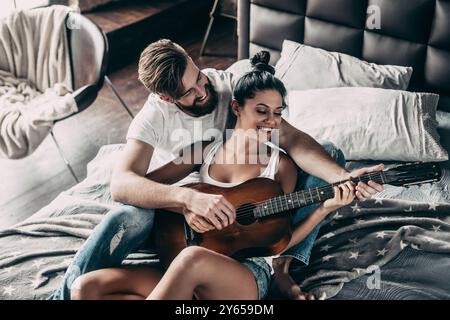 Modern couple. Top view of handsome young bearded man teaching his girlfriend to play guitar while both sitting in bed at home together Stock Photo