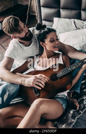Beautiful couple playing guitar. Handsome young bearded man teaching his girlfriend to play guitar while both sitting in bed at home together Stock Photo