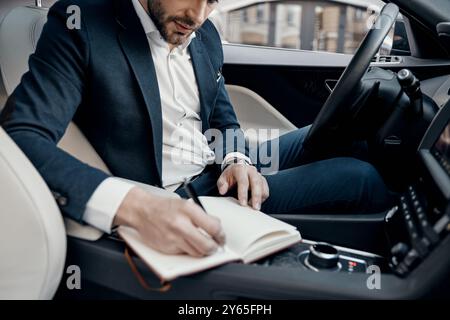 Business is his life. Close up of young man in full suit writing something down in personal organizer while sitting inside of the car Stock Photo
