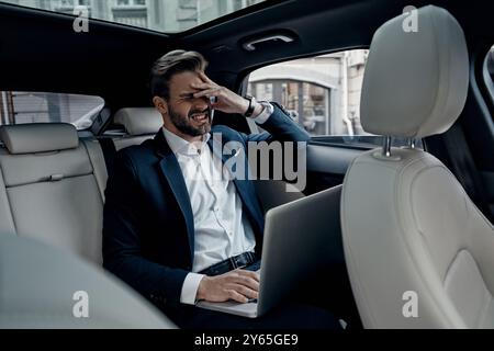 Oh no! Frustrated young man in full suit working using laptop and covering face with hand while sitting in the car Stock Photo