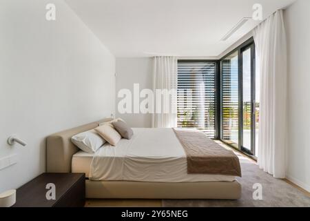 A contemporary bedroom featuring minimalist design in a luxury Mediterranean villa, with sunlight streaming through large windows with blinds Stock Photo