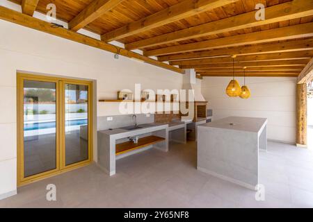 Modern Ibizan villa kitchen features white minimalist design with wooden beams, a central island, and Mediterranean styled pendant lights Stock Photo