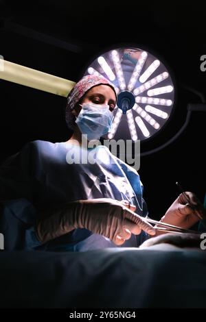 A focused female surgeon performs a procedure in an operating room, illuminated by a large surgical light, capturing the intensity and precision of me Stock Photo