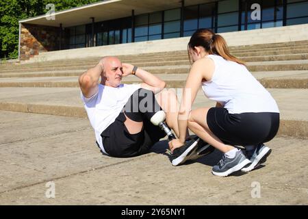 A middle-aged man with an amputated leg, sporting a prosthetic, exercises on steps outdoors under the assistance of his young adult daughter Stock Photo
