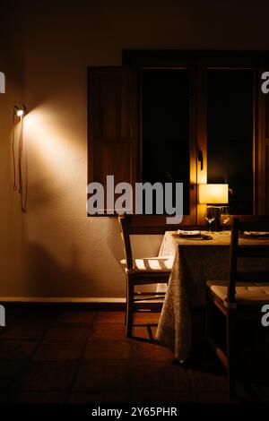 A warmly lit, intimate dining setting inside a rustic room, featuring an elegantly set table with two glasses and a quaint window providing soft eveni Stock Photo