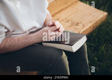 Cropped image of an unrecognizable man sitting on a wooden bench, holding and reading a Bible outdoors with a scenic natural backdrop. Stock Photo