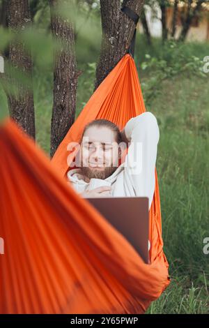 A serene image capturing a young man lying comfortably in an orange hammock between trees, using a laptop in a tranquil outdoor woodland setting Stock Photo