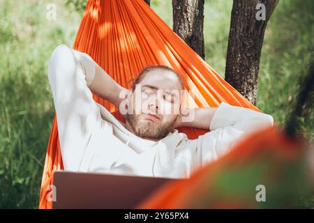 A serene image of a young man peacefully sleeping in an orange hammock tied between trees, set against a lush green backdrop, perfect for themes of re Stock Photo