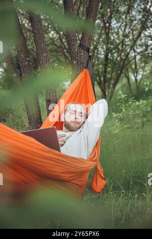 Young man enjoys tranquility while lying in an orange hammock with his laptop, nestled among green trees in a serene forest setting Stock Photo