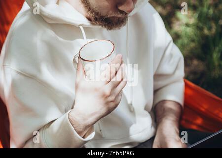 A peaceful setting capturing a young man lounging in a hammock, holding a mug of coffee, dressed in a white hoodie, illustrating relaxation in a natur Stock Photo