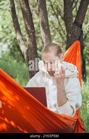 A young man relaxes in an orange hammock between trees, focusing intently on his laptop while drinking coffee in a serene natural setting Stock Photo