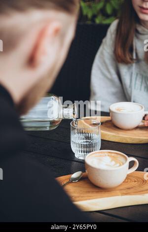 Cropped unrecognizable couple having coffee at a cafe, with a focus on a man pouring water into a glass and a woman with a cup of cappuccino. The scen Stock Photo
