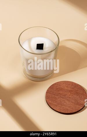 A minimalist depiction of a scented white candle in a transparent glass, accompanied by a round cork coaster on a beige surface under soft sunlight Stock Photo