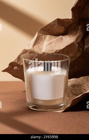 Close-up of an elegant white candle placed in a transparent glass The candle is set against a textured brown paper backdrop with a subtle play of ligh Stock Photo