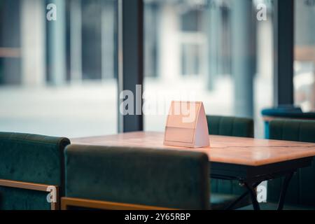 Contemporary cafe corner with a stylish wooden table featuring a geometric menu holder, set against a blurred urban backdrop through large windows. Stock Photo