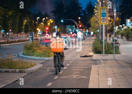 Back view of unrecognizable food delivery cyclist navigates the busy twilight streets of Madrid near AZCA financial district, showcasing modern urban Stock Photo