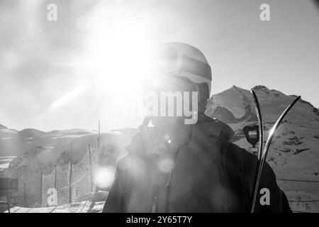 A skier, silhouetted against a sunlit snowy landscape, stands with skis beside a mountain slope, embodying a serene winter scene Stock Photo